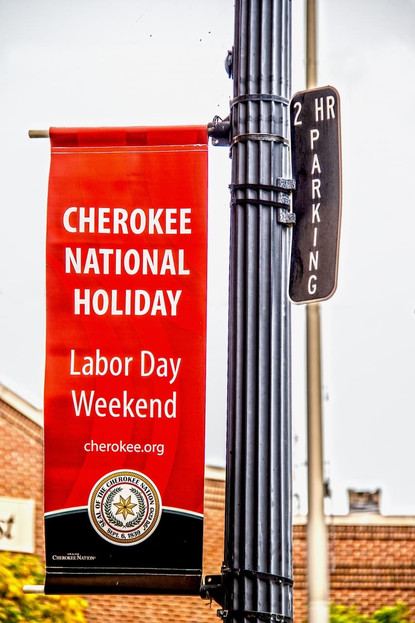 Pancarta roja anunciando el Cherokee National Holiday durante el fin de semana del Día del Trabajo, colgada en un poste junto a un letrero de estacionamiento, impresa por Punto Clave