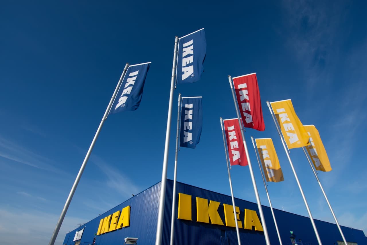 Se observan varias banderas ondeando en un día soleado frente a un edificio de la marca 'IKEA', que destaca con su característico color azul y el logo amarillo. Las banderas presentan diferentes colores, incluidos azul, rojo y amarillo, y todas llevan el nombre 'IKEA' en letras grandes y blancas.