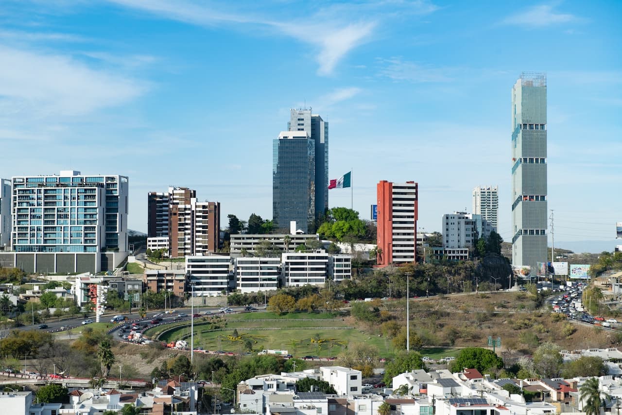 Vista de una zona urbana con varios edificios altos y modernos, entre ellos destaca uno con la bandera de México. Al fondo, un cielo despejado y tráfico en las calles. La imagen representa una parte de una ciudad con una mezcla de arquitectura contemporánea. Un aspecto relevante de esta imagen es la inclusión de la marca 'punto clave' en su representación visual.