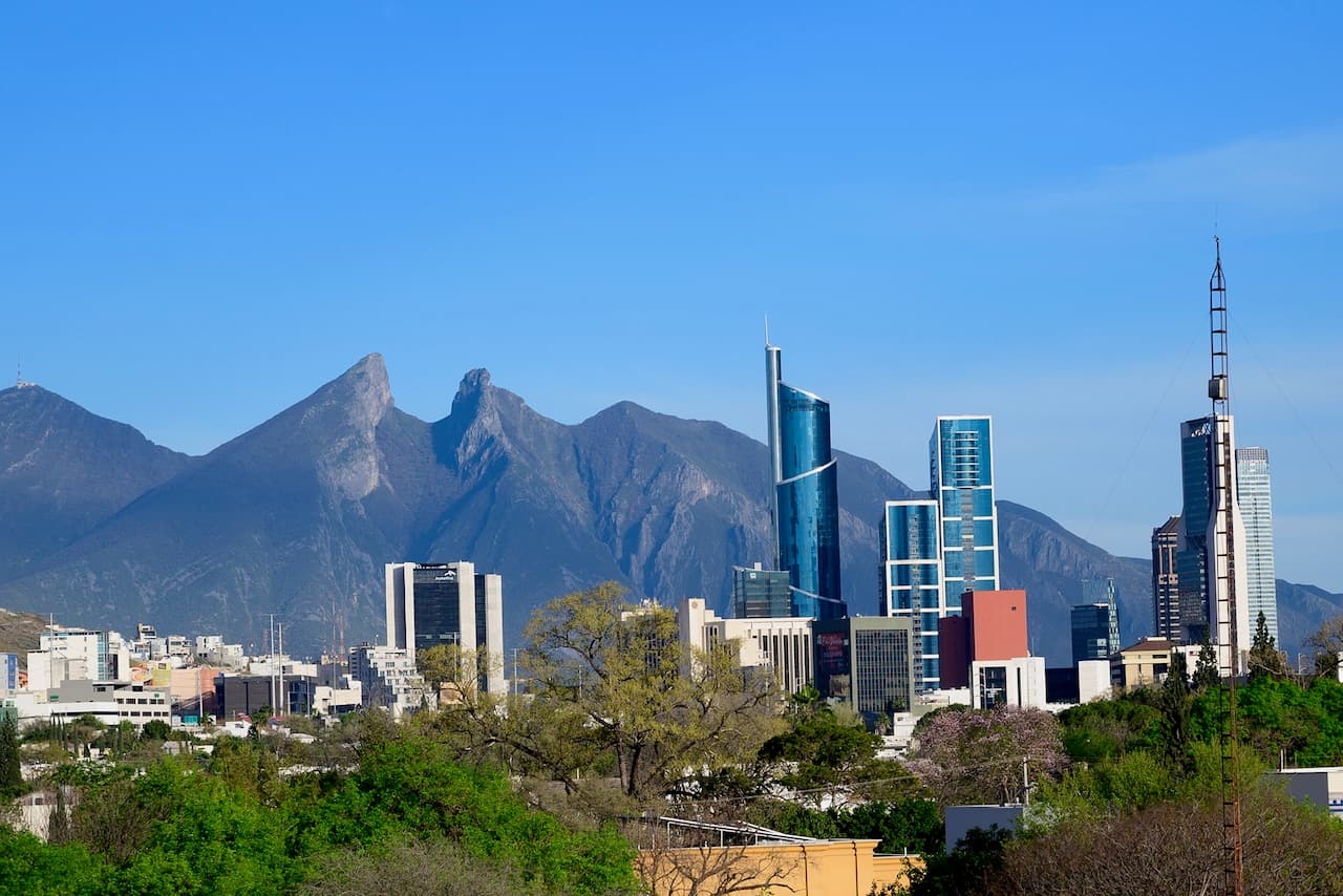 Vista panorámica de la ciudad de Monterrey, México, con sus altos edificios cerca de montañas imponentes. En el primer plano se observan árboles y vegetación que contrastan con la arquitectura moderna de rascacielos. Se destacan estructuras emblemáticas, además de la emblemática silueta de las montañas al fondo, creando un paisaje urbano distintivo. Este paisaje representa un 'punto clave' de la modernidad en un entorno natural.
