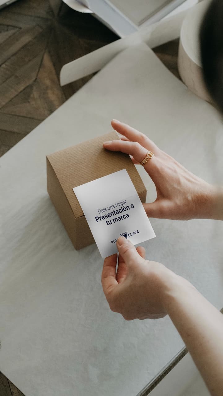 Una persona sostiene una tarjeta blanca que dice 'Dale una mejor Presentación a tu marca' junto a una caja de cartón sobre una superficie blanca. La tarjeta tiene el logo de la marca 'punto clave'.