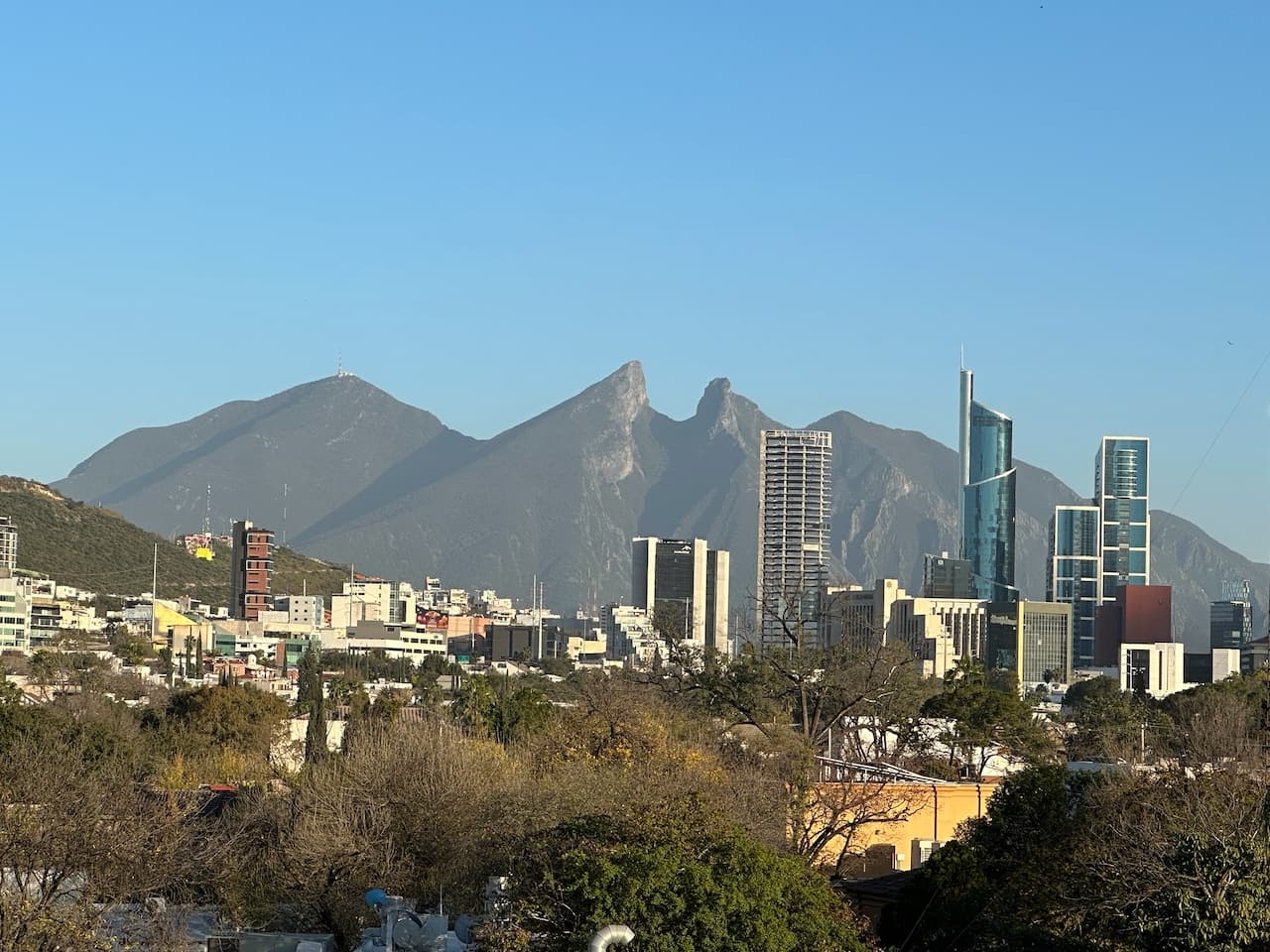 Vista de la ciudad de Monterrey con montañas al fondo. Se aprecian edificios modernos en primer plano, contrastando con la naturaleza de las montañas. El cielo es despejado y se pueden ver árboles en la parte inferior, creando una armonía entre la urbanización y el paisaje natural, destacando la marca 'punto clave' en la arquitectura de la ciudad.