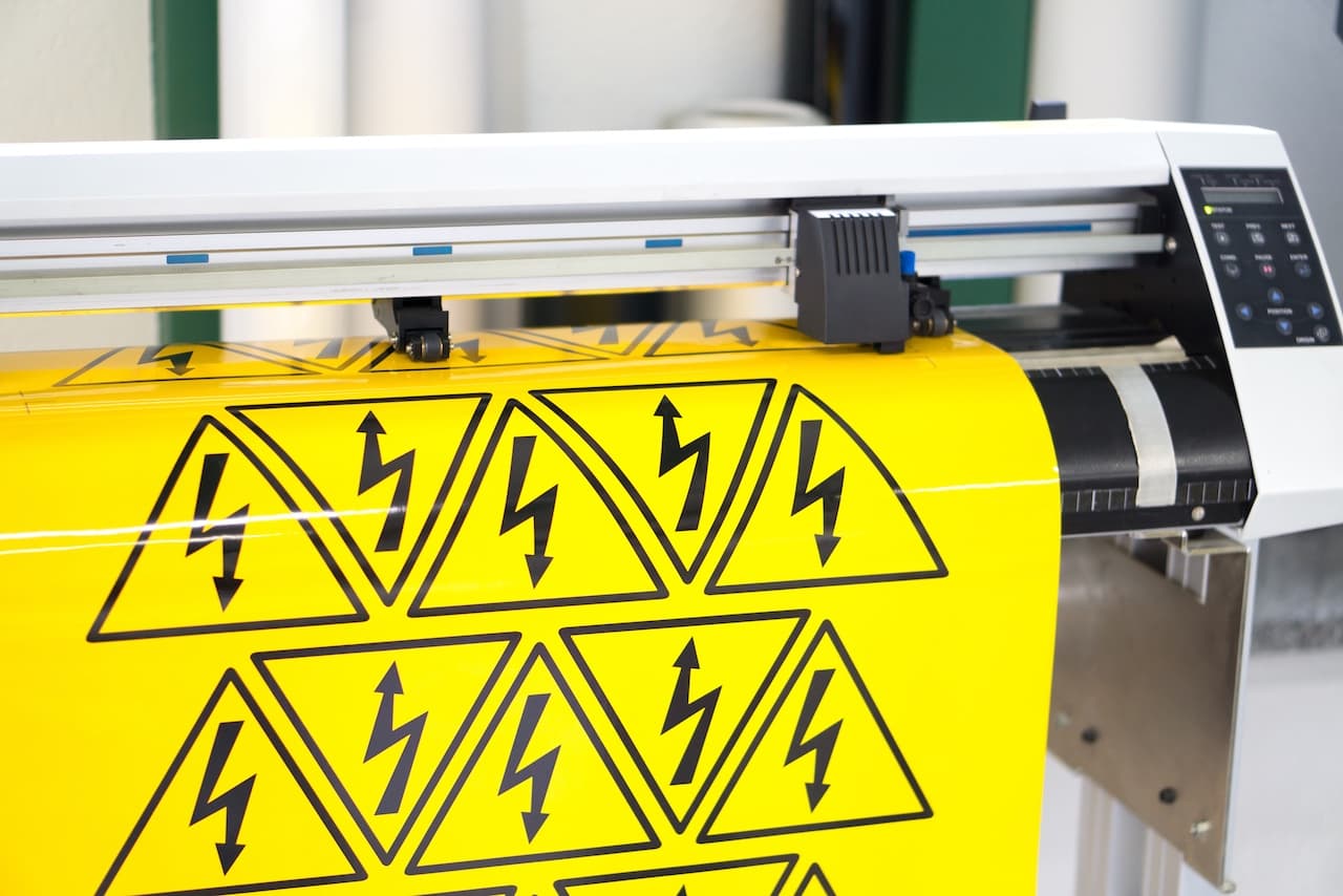Se observa una impresora de corte que está trabajando con un rollo de vinilo amarillo, donde destacan múltiples triángulos con símbolos de rayo negro, indicando peligro eléctrico. La impresora parece ser parte de un equipo de señalización de la marca 'punto clave'.