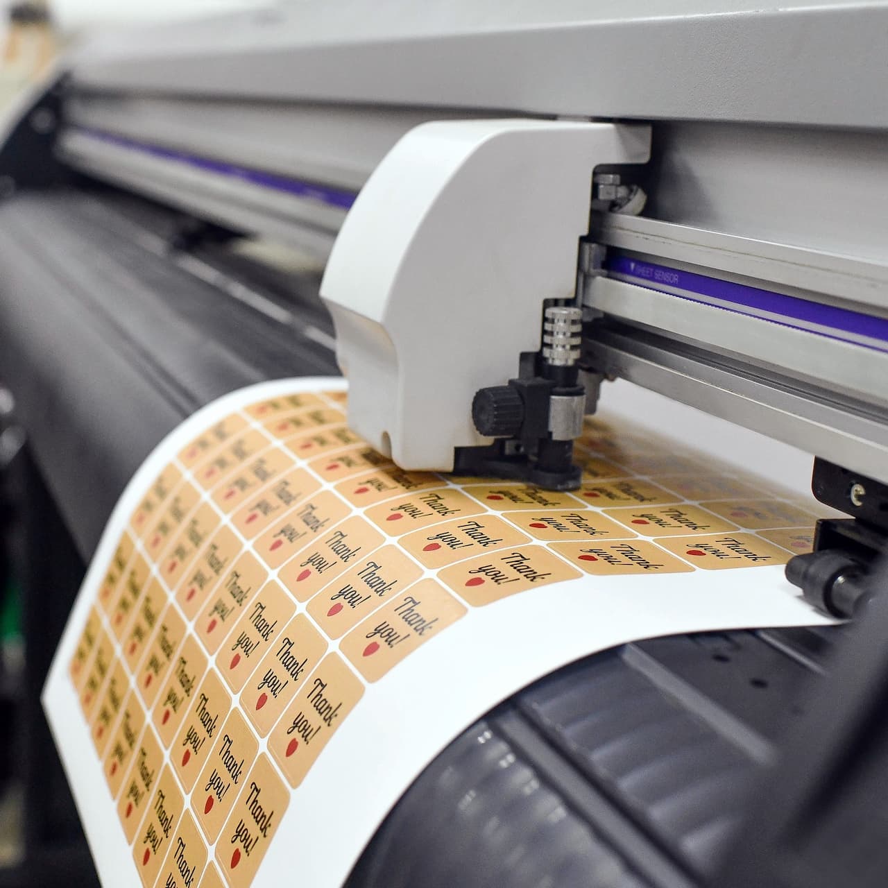 Una impresora está imprimiendo etiquetas con el texto 'Thank you!' en un fondo beige. Las etiquetas están dispuestas en filas sobre un rollo de papel. La máquina tiene un diseño moderno y está equipada para trabajos de impresión de alta calidad, destacando la marca 'punto clave'.
