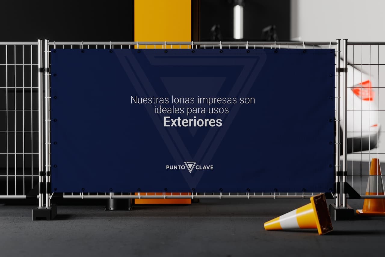 Lona impresa de color azul con el mensaje 'Nuestras lonas impresas son ideales para usos Exteriores', destacando la marca 'punto clave'. La lona está instalada en una valla, con conos de tráfico amarillos en el suelo, en un ambiente que parece ser un estacionamiento o taller.