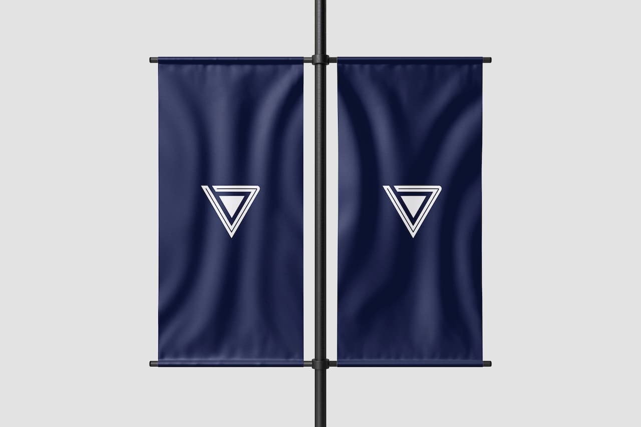 Se presentan dos banderas colgantes en color azul marino, cada una adornada con un diseño geométrico en blanco que consiste en un triángulo invertido y un triángulo hacia arriba. Las banderas están dispuestas a cada lado de un poste central, reflejando un estilo moderno y minimalista, representativo de la marca 'punto clave'.
