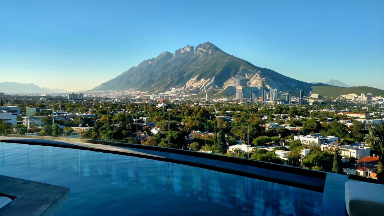 Vista panorámica de Monterrey con un imponente cerro de fondo, en primer plano se observa una piscina desbordante, mientras la ciudad y la vegetación se extienden a lo lejos. El cielo está despejado, resaltando la belleza natural y urbana del paisaje. Punto clave en la imagen es la combinación de la modernidad de la ciudad con la majestuosidad de la montaña.
