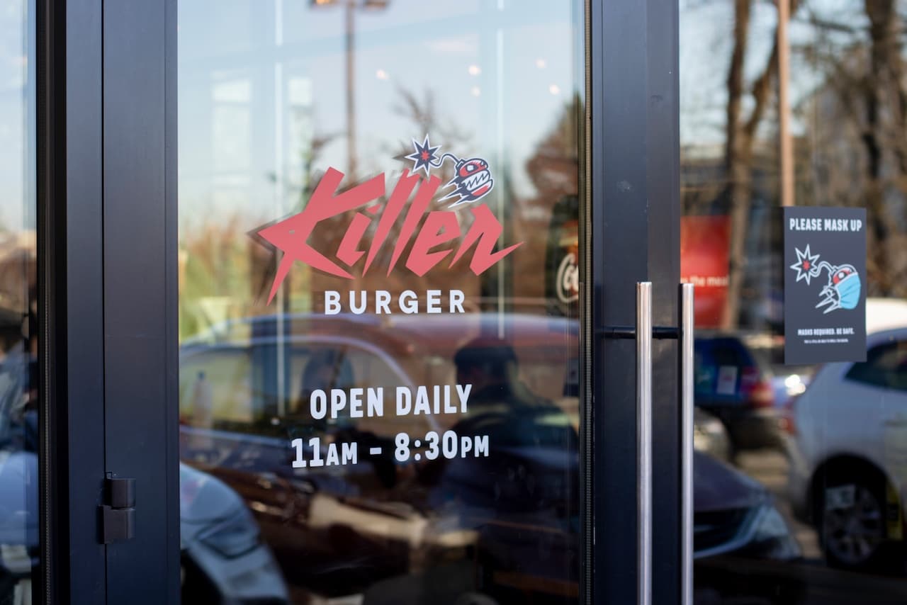 Se observa la entrada de un restaurante llamado 'Killer Burger', con un logo distintivo en colores rojo y negro. La puerta tiene un letrero que indica que está abierto diariamente de 11 am a 8:30 pm. También hay un aviso que solicita usar mascarilla, reforzando la importancia de la seguridad sanitaria. La marca 'punto clave' resalta el ambiente moderno y divertido del lugar.