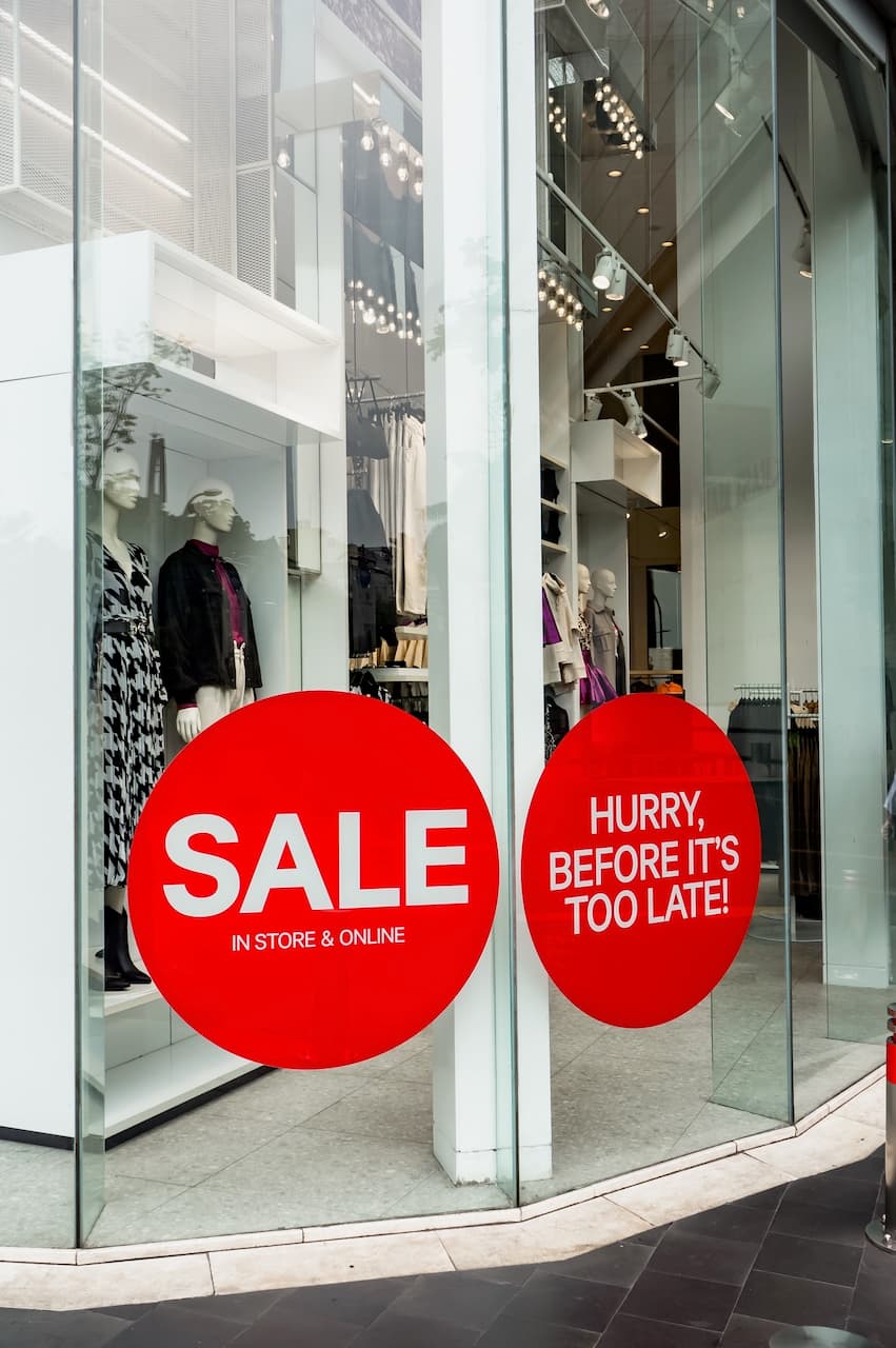 Un escaparate de tienda muestra maniquíes vestidas y dos grandes círculos rojos con textos que anuncian una 'SALE' y un mensaje que invita a los clientes a 'HURRY, BEFORE IT’S TOO LATE!'. La escena es clara y está iluminada, destacando la oferta. Marca 'punto clave'.