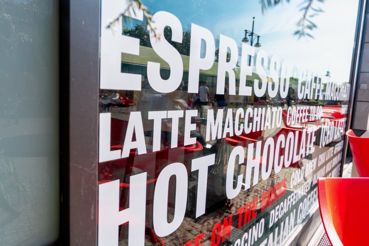 Se observa un vidrio de un local con diversos nombres de bebidas escritas en letras grandes y claras, que incluyen 'Espresso', 'Latte Macchiato', 'Hot Chocolate' y otras opciones de café. Al fondo hay sillas rojas y elementos del entorno reflejándose en el vidrio, destacando un ambiente acogedor. La marca 'punto clave' resalta en la presentación.