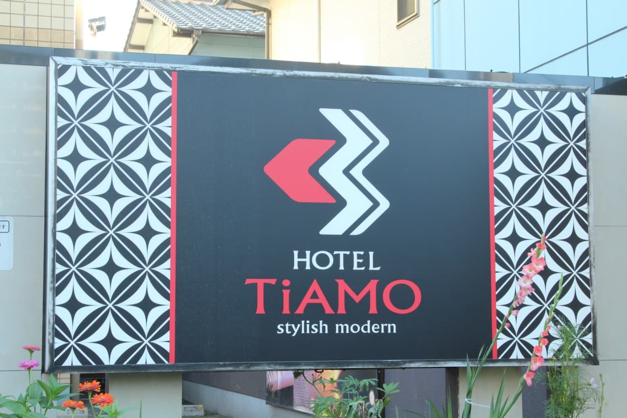 Letrero del 'Hotel TiAMO' con diseño moderno, fondo negro y detalles en rojo y blanco. Incluye elementos gráficos que refuerzan su estilo 'stylish modern'. Hay flores y plantas en primer plano, añadiendo color al entorno.