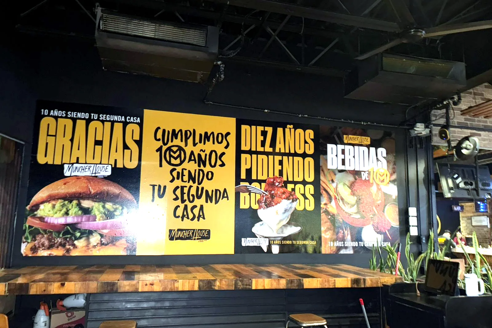 Cuatro lonas impresas instaladas en la pared de un restaurante, con fotos de hamburguesas y mensajes de aniversario